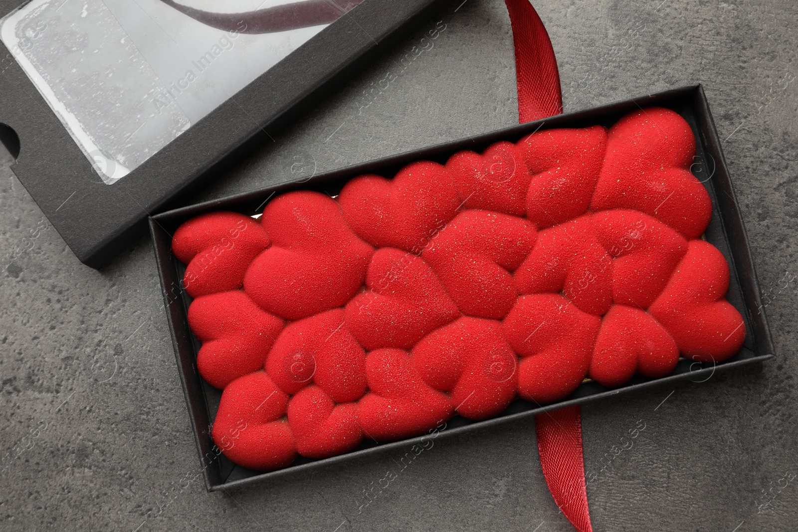 Photo of Chocolate bar decorated with hearts in box and red ribbon on grey table, flat lay