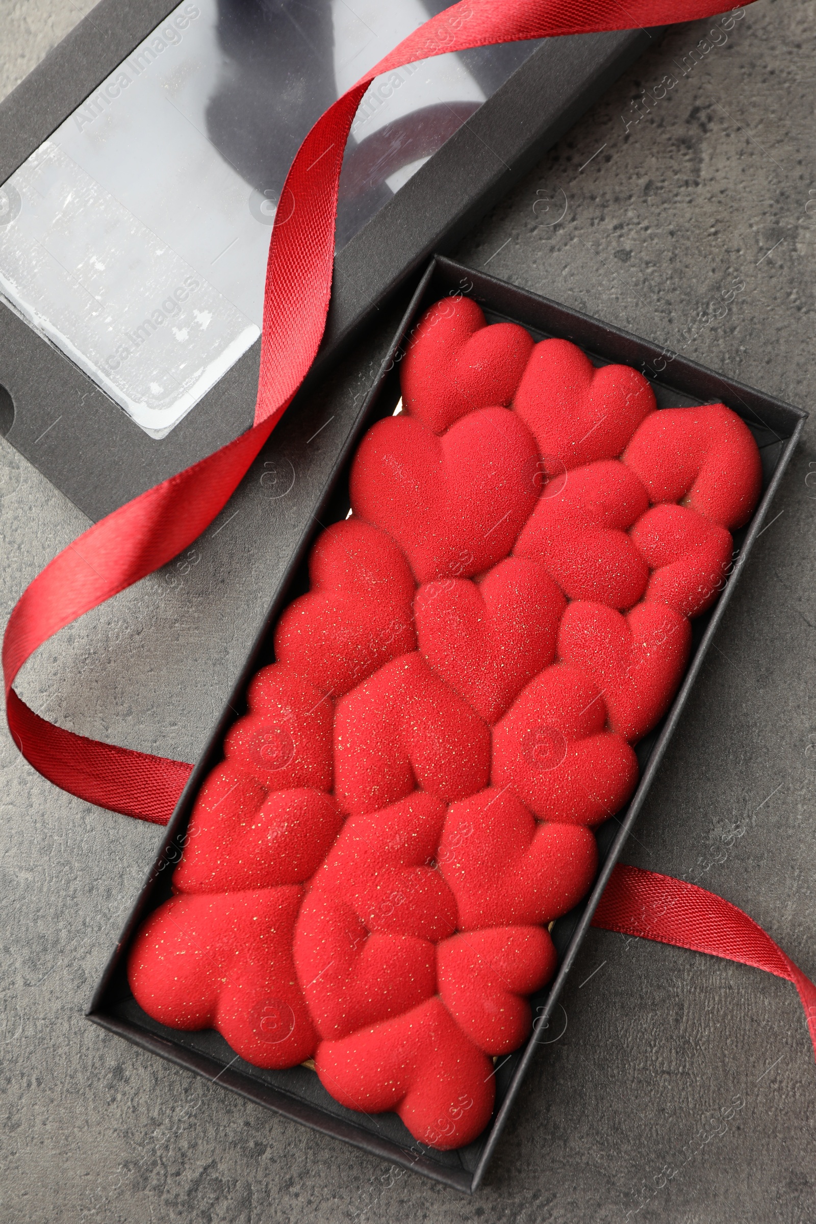 Photo of Chocolate bar decorated with hearts in box and red ribbon on grey table, flat lay