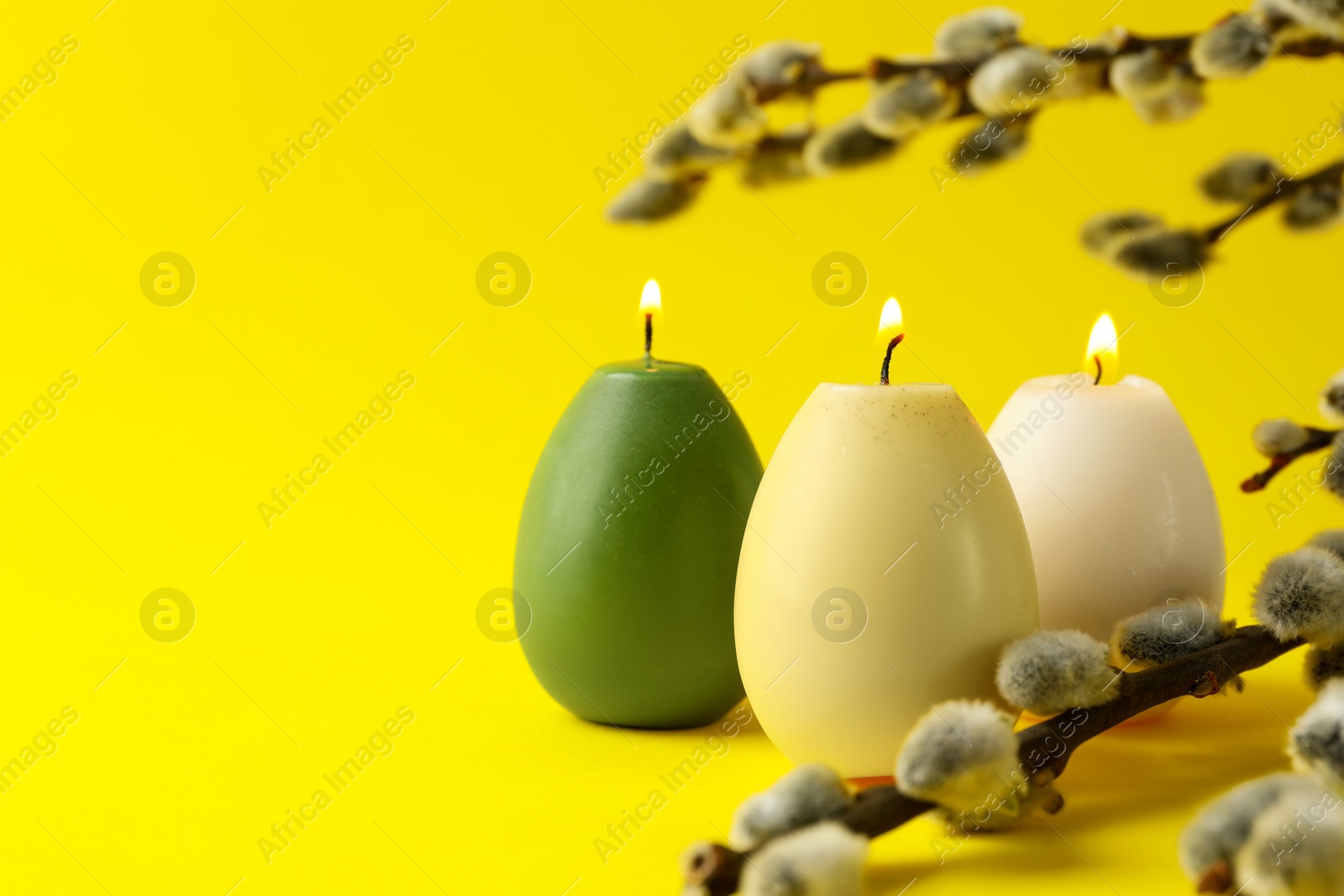 Photo of Beautiful burning egg shaped candles and pussy willow on yellow background, closeup with space for text. Easter decor