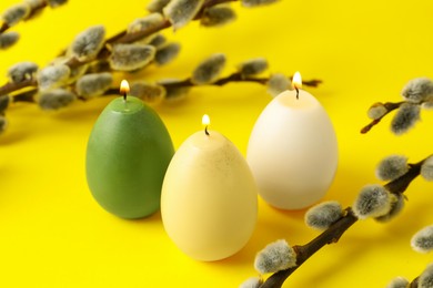 Photo of Beautiful burning egg shaped candles and pussy willow on yellow background, closeup. Easter decor