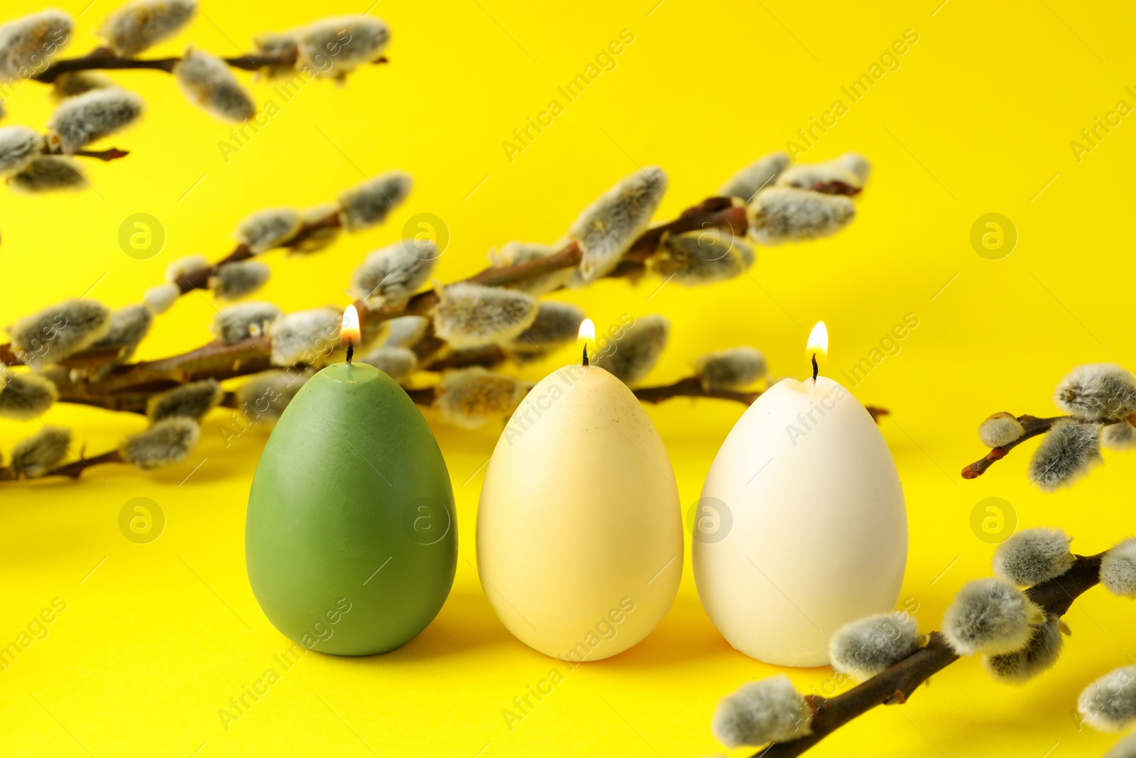 Photo of Beautiful burning egg shaped candles and pussy willow on yellow background. Easter decor