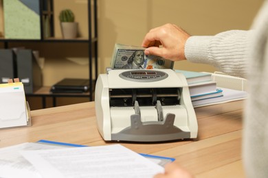 Photo of Man putting money into banknote counter at wooden table indoors, closeup