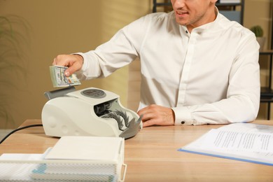 Photo of Man putting money into banknote counter at wooden table indoors, closeup