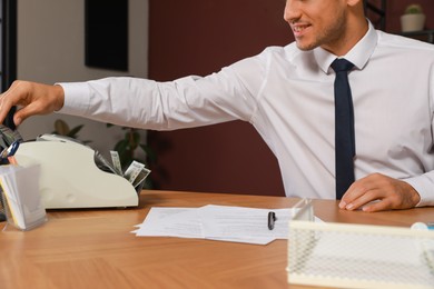 Photo of Man putting money into banknote counter at wooden table indoors, closeup