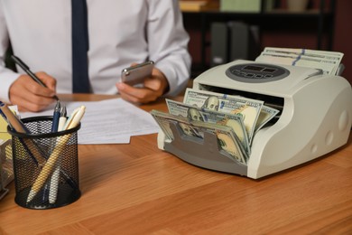 Photo of Modern banknote counter with money and blurred view of man working at wooden table indoors