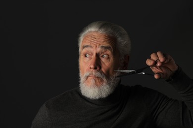 Photo of Senior man trimming beard with scissors on dark grey background