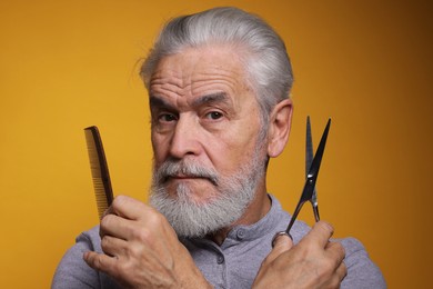 Photo of Bearded senior man holding comb and scissors on orange background