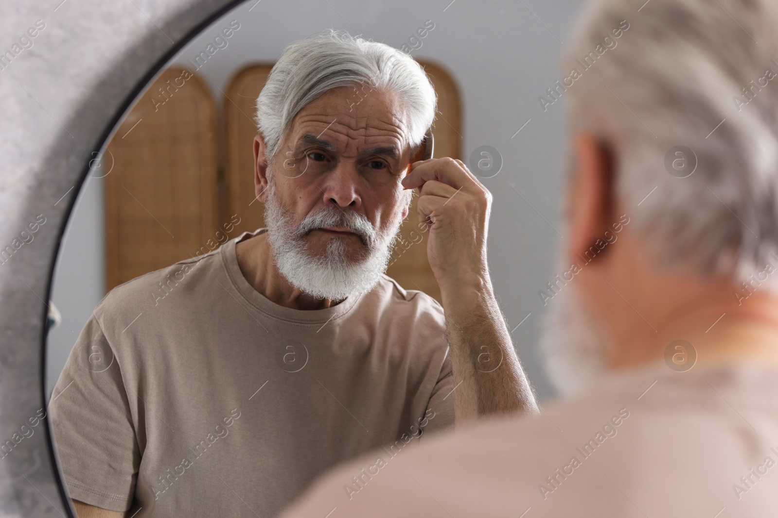 Photo of Bearded senior man combing hair near mirror in bathroom