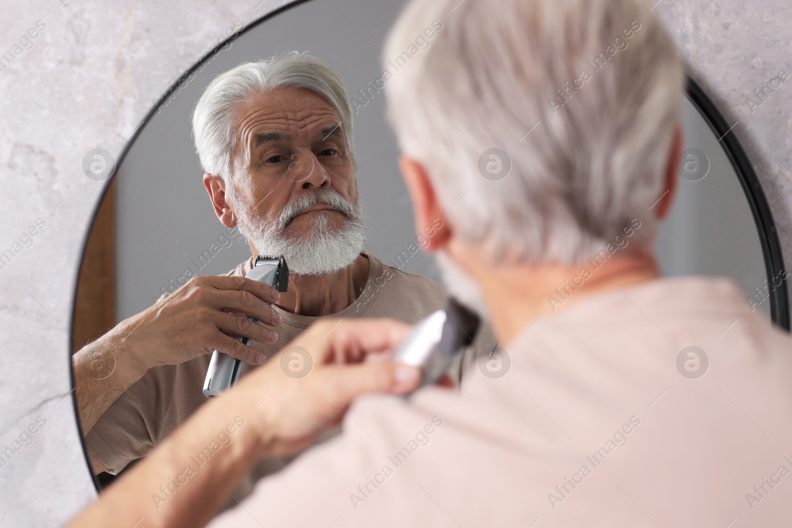 Photo of Senior man trimming beard near mirror in bathroom