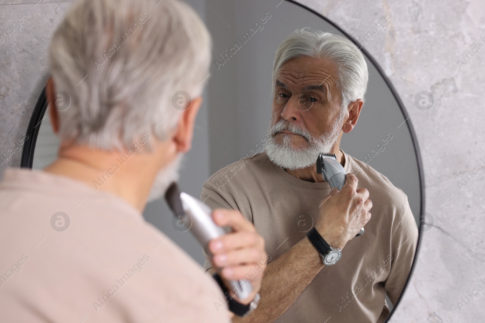 Photo of Senior man trimming beard near mirror in bathroom