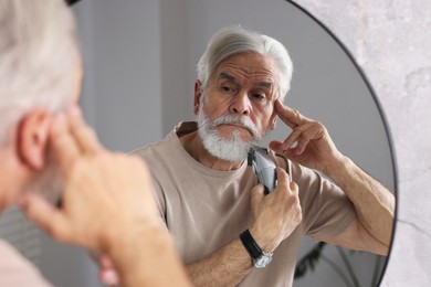 Photo of Senior man trimming beard near mirror in bathroom