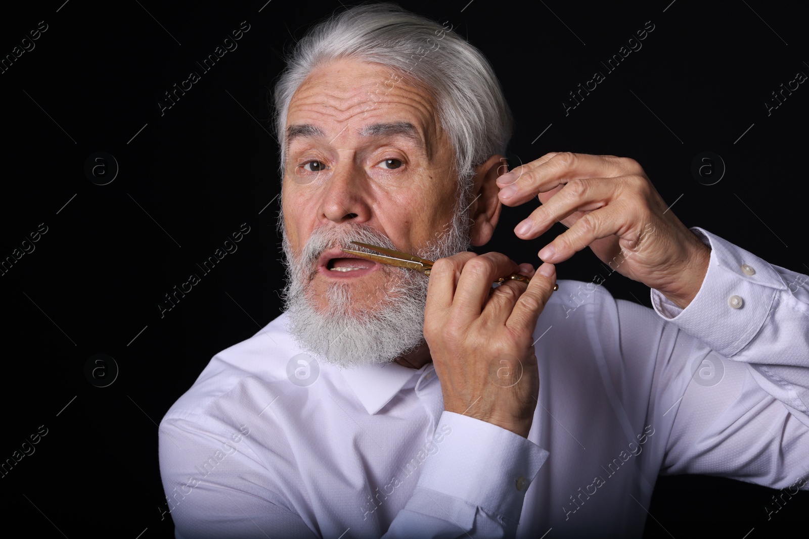 Photo of Senior man trimming beard with scissors on black background