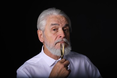 Photo of Senior man trimming beard with scissors on black background