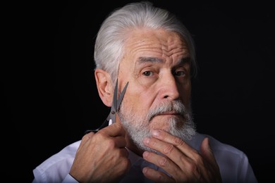 Photo of Senior man trimming beard with scissors on black background