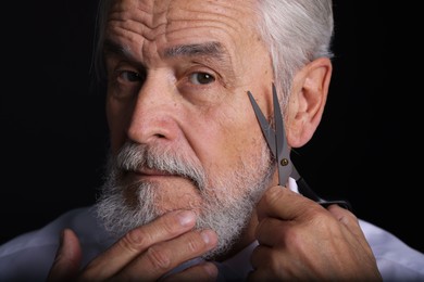 Senior man trimming beard with scissors on black background