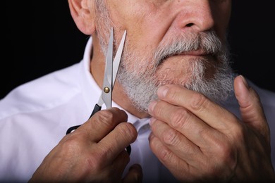 Photo of Senior man trimming beard with scissors on black background, closeup