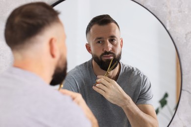 Photo of Man trimming beard with scissors near mirror in bathroom