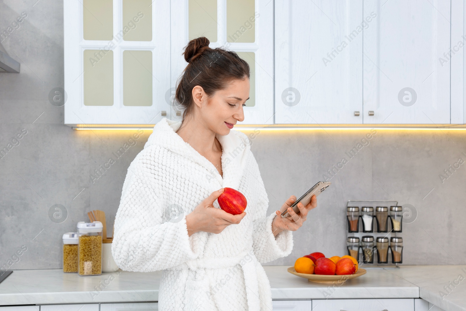 Photo of Beautiful woman with apple using smartphone after spa procedure in kitchen