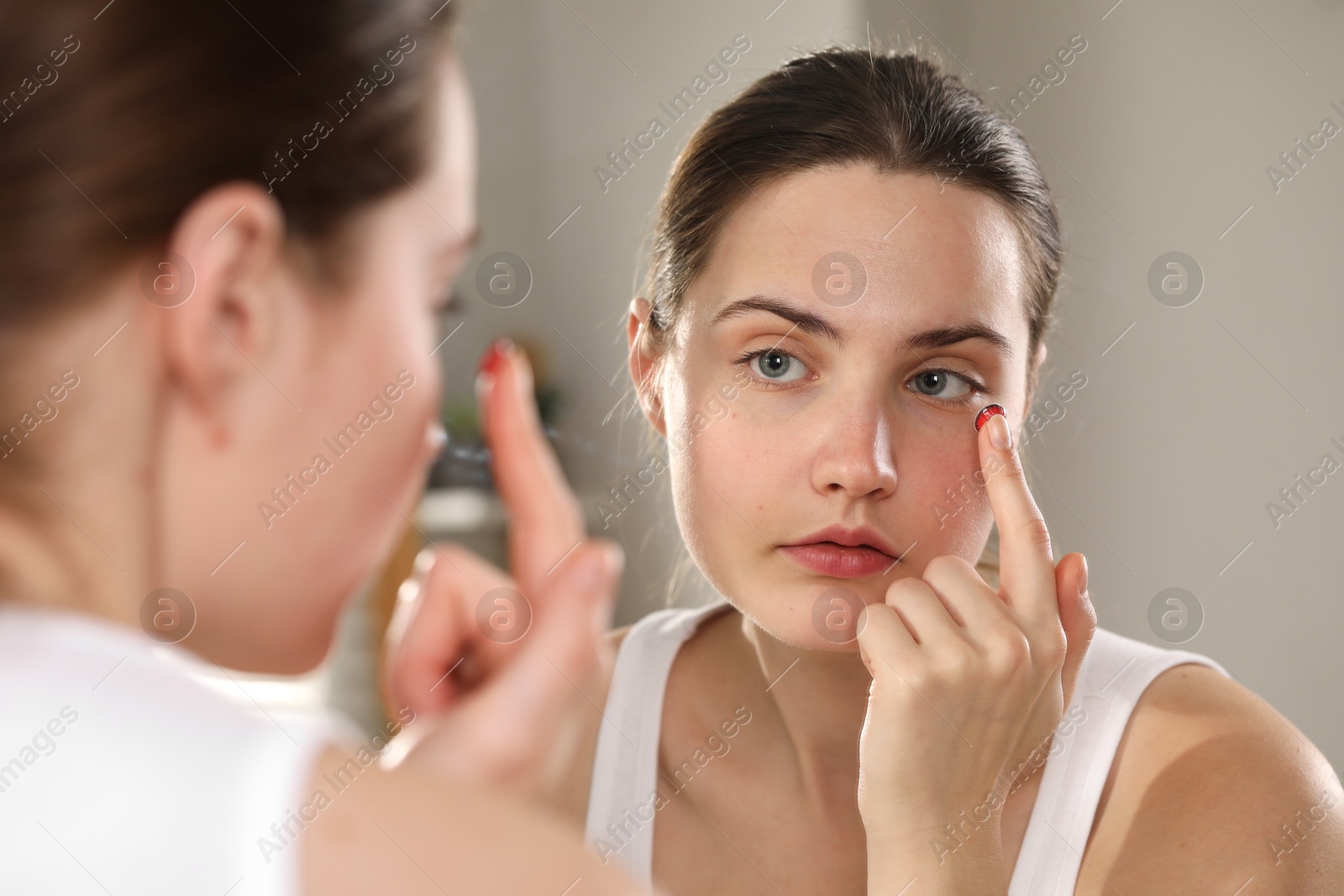 Photo of Young woman putting in red color contact lens near mirror indoors