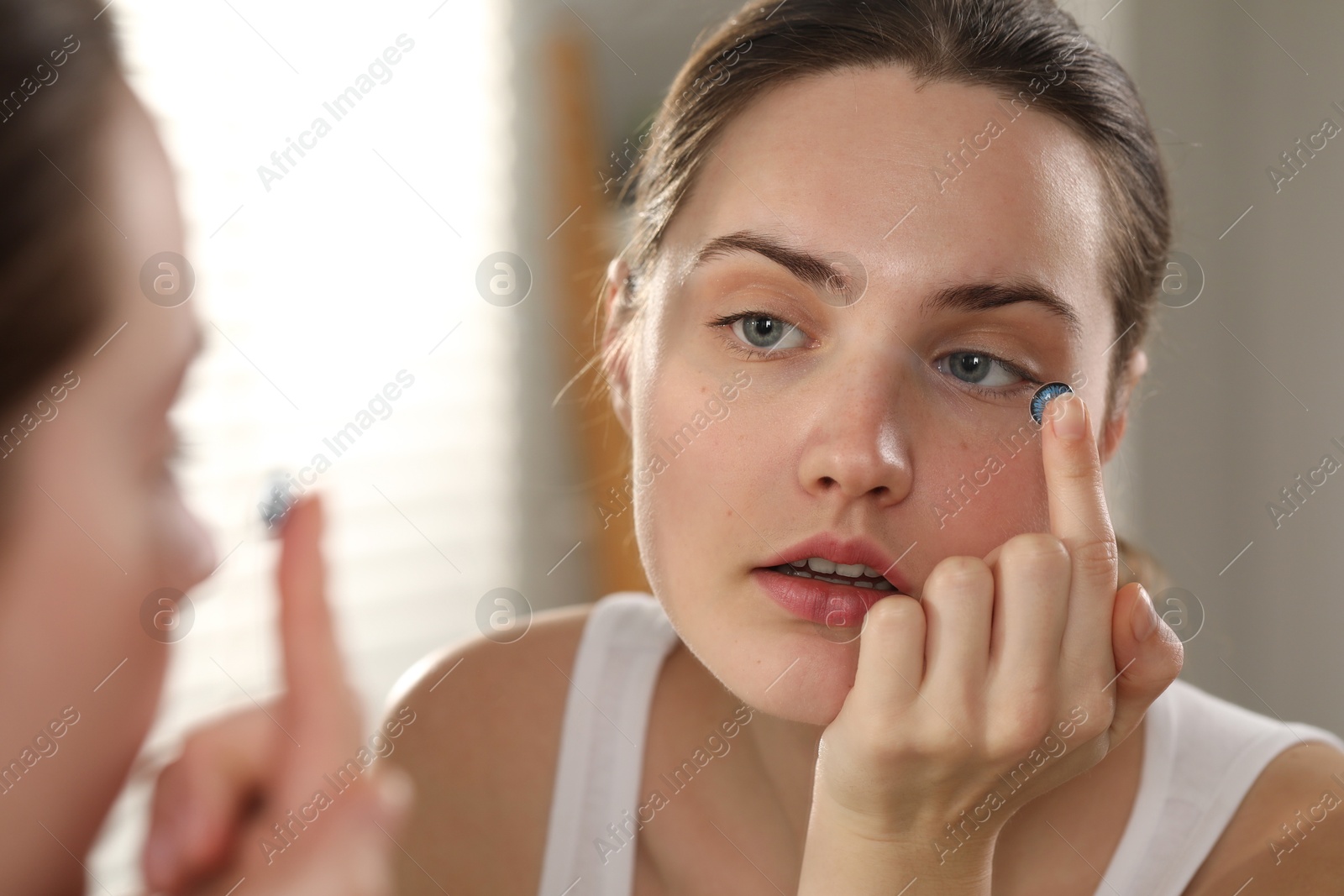 Photo of Young woman putting in blue color contact lens near mirror indoors, closeup