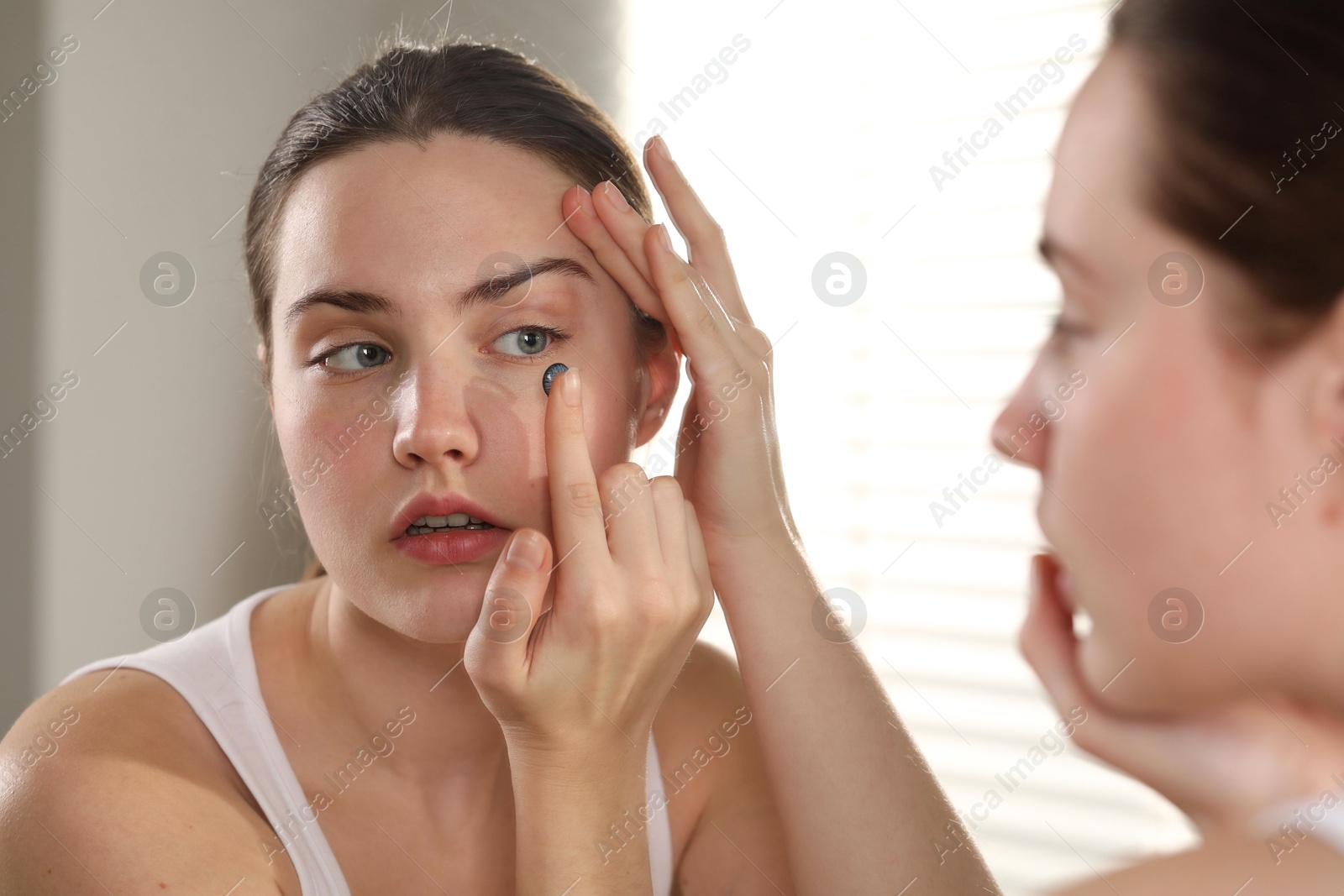 Photo of Young woman putting in blue color contact lens near mirror indoors