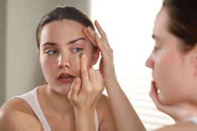 Photo of Young woman putting in blue color contact lens near mirror indoors