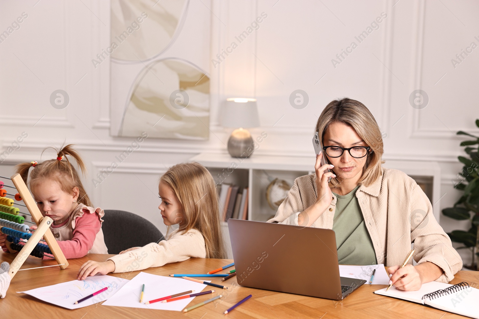 Photo of Work-family balance. Single mother talking by smartphone and her children at wooden table indoors