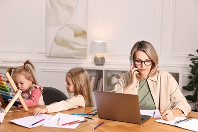 Photo of Work-family balance. Single mother talking by smartphone and her children at wooden table indoors