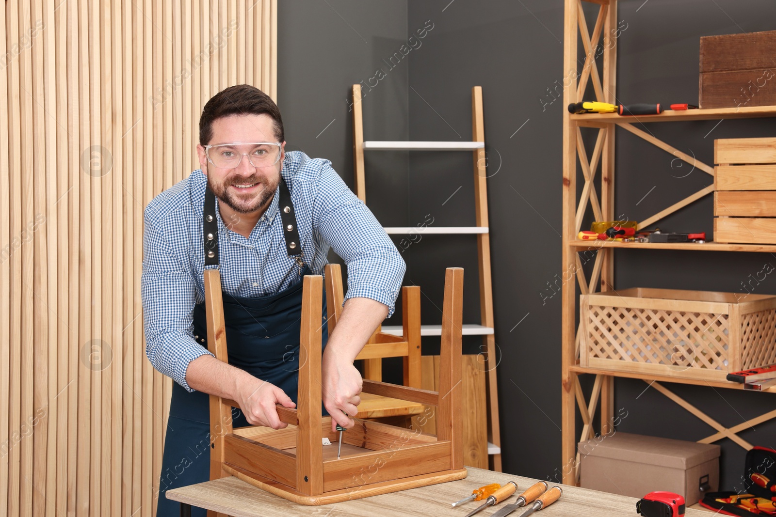 Photo of Relaxing hobby. Smiling man assembling wooden chair with screwdriver at table in workshop