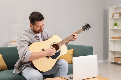 Photo of Relaxing hobby. Man playing guitar with online tutorial by laptop on sofa at home