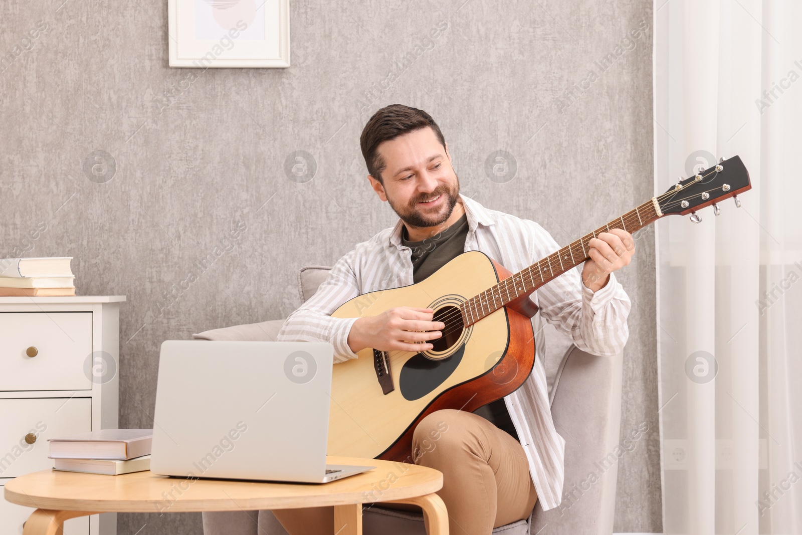 Photo of Relaxing hobby. Smiling man playing guitar with online tutorial by laptop on armchair at home