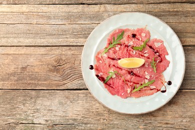 Photo of Delicious beef carpaccio with arugula, cheese, balsamic vinegar and lemon on wooden table, top view. Space for text