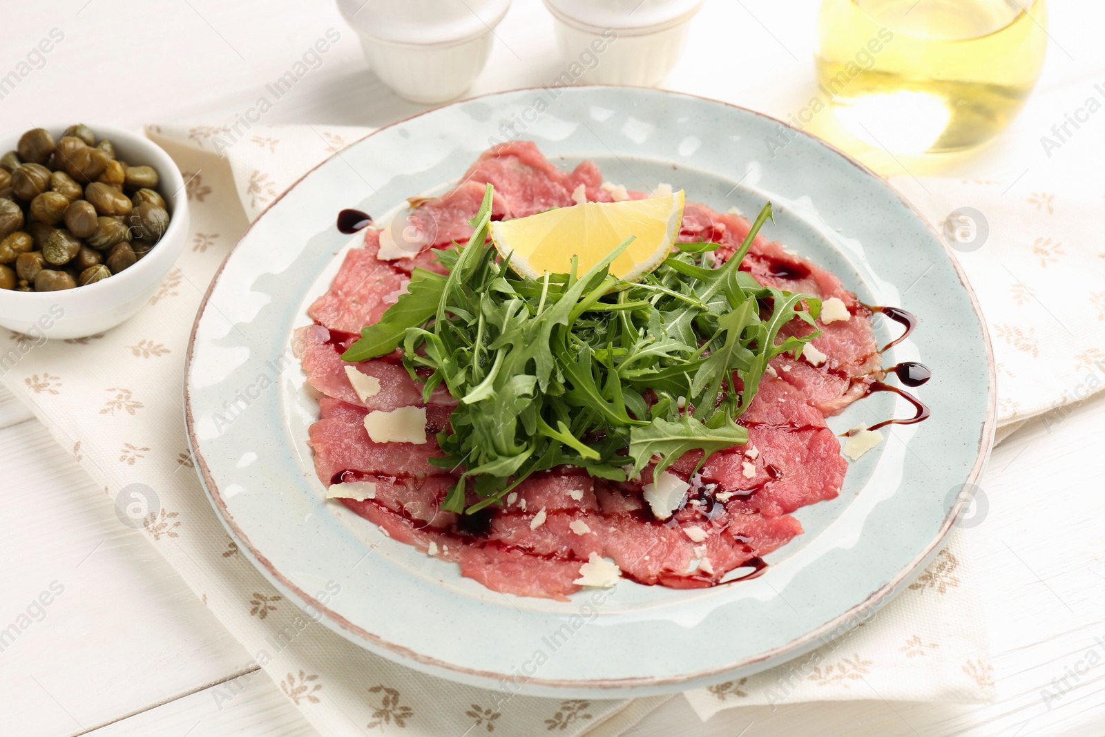 Photo of Delicious beef carpaccio with arugula, cheese, balsamic vinegar and lemon on white wooden table, closeup