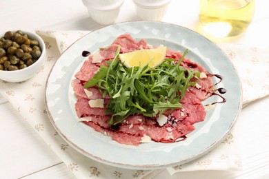 Photo of Delicious beef carpaccio with arugula, cheese, balsamic vinegar and lemon on white wooden table, closeup