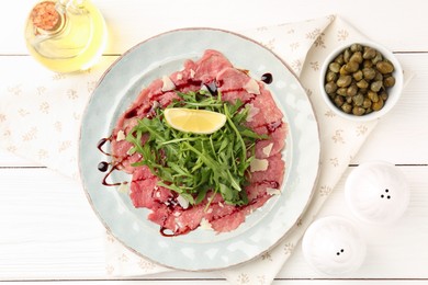 Photo of Delicious beef carpaccio with arugula, lemon, capers and oil on white wooden table, flat lay