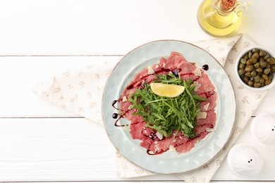 Photo of Delicious beef carpaccio with arugula, lemon, capers and oil on white wooden table, flat lay. Space for text