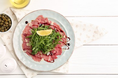 Photo of Delicious beef carpaccio with arugula, lemon, capers and oil on white wooden table, flat lay. Space for text