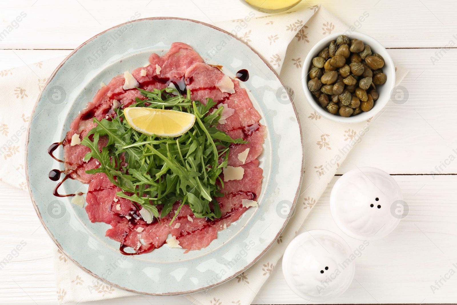 Photo of Delicious beef carpaccio with arugula, cheese, lemon and capers on white wooden table, flat lay