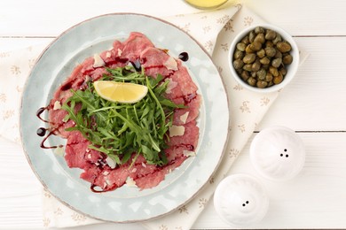 Photo of Delicious beef carpaccio with arugula, cheese, lemon and capers on white wooden table, flat lay