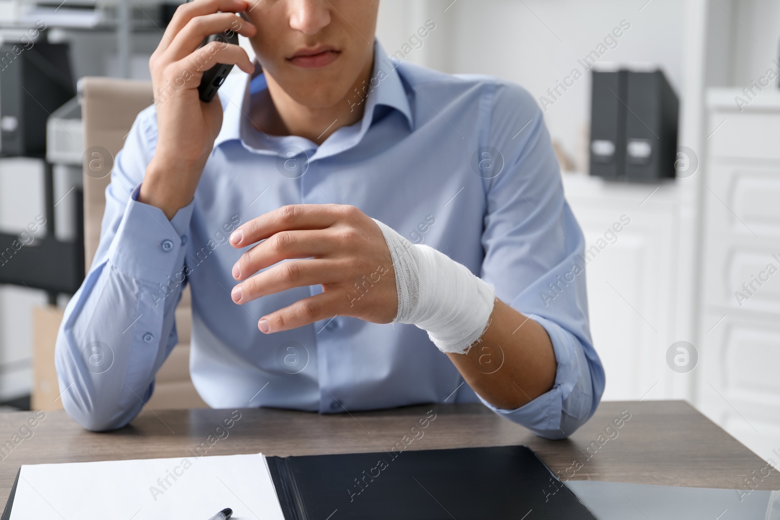 Photo of Man with wrist wrapped in medical bandage talking on smartphone at table indoors, closeup