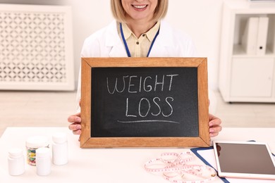 Photo of Happy nutritionist holding small blackboard with words Weight loss at table in clinic, closeup