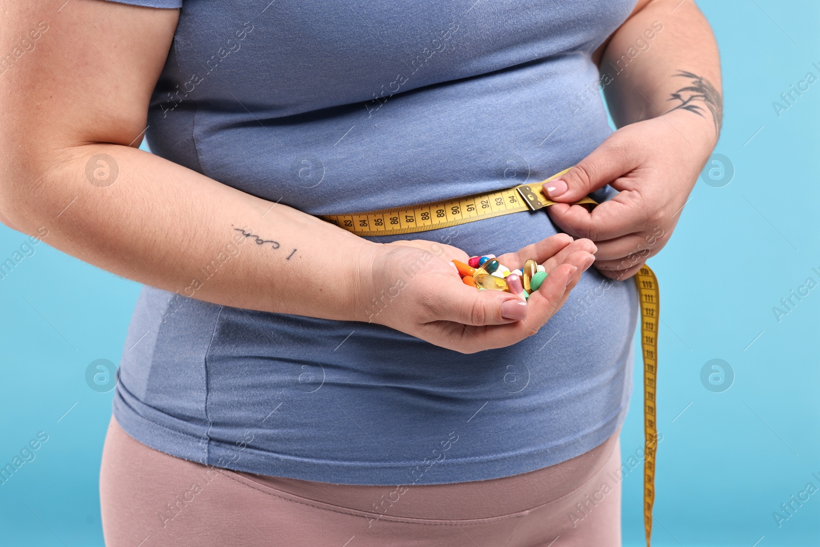 Photo of Plus size woman measuring waist with tape and holding pile of weight loss supplements on light blue background, closeup