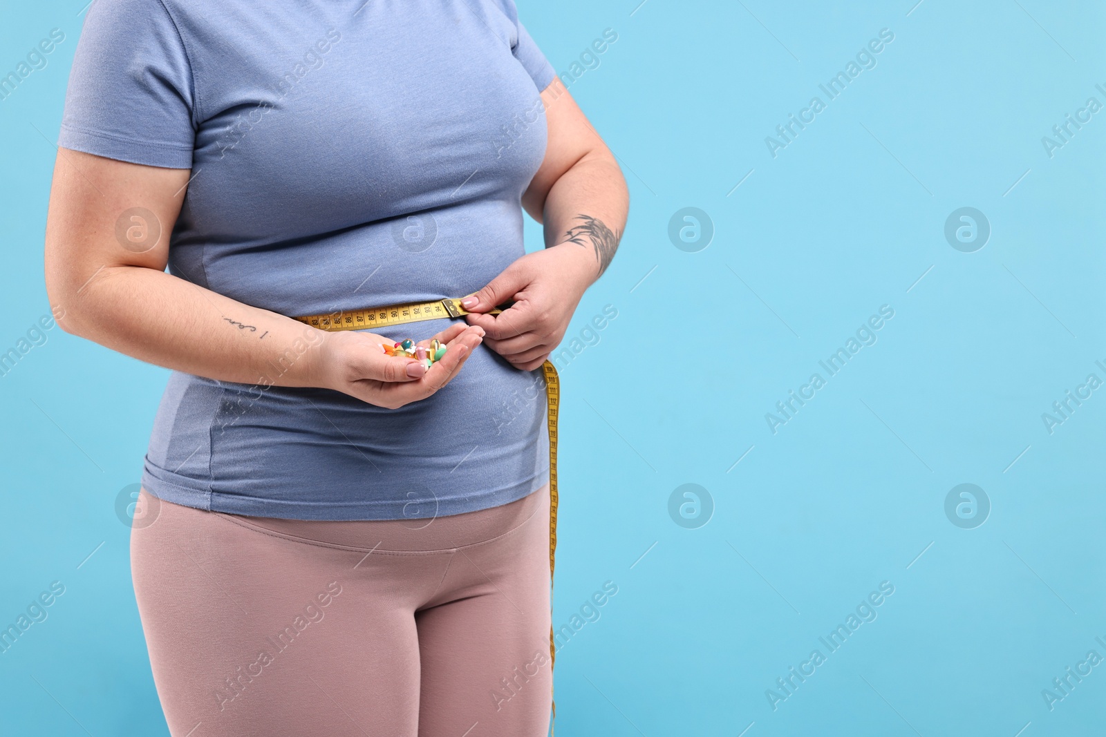 Photo of Plus size woman measuring waist with tape and holding pile of weight loss supplements on light blue background, closeup. Space for text