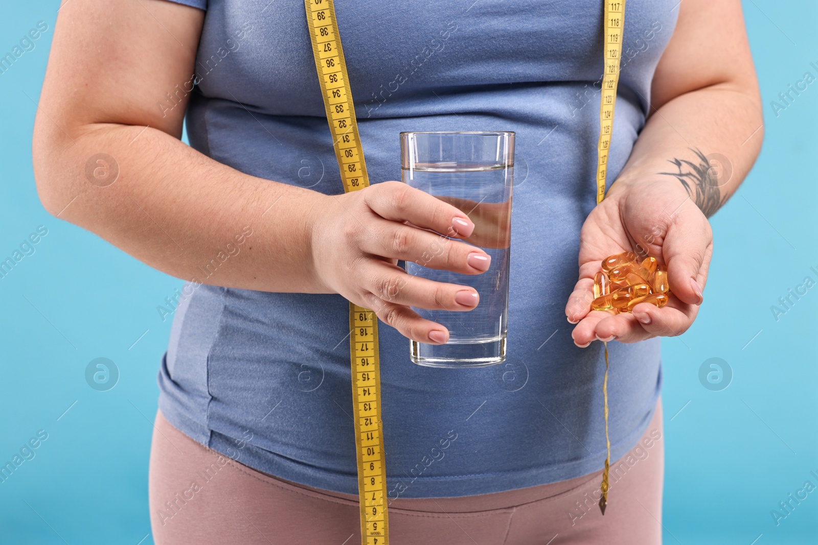 Photo of Plus size woman holding pile of weight loss supplements and glass with water on light blue background, closeup