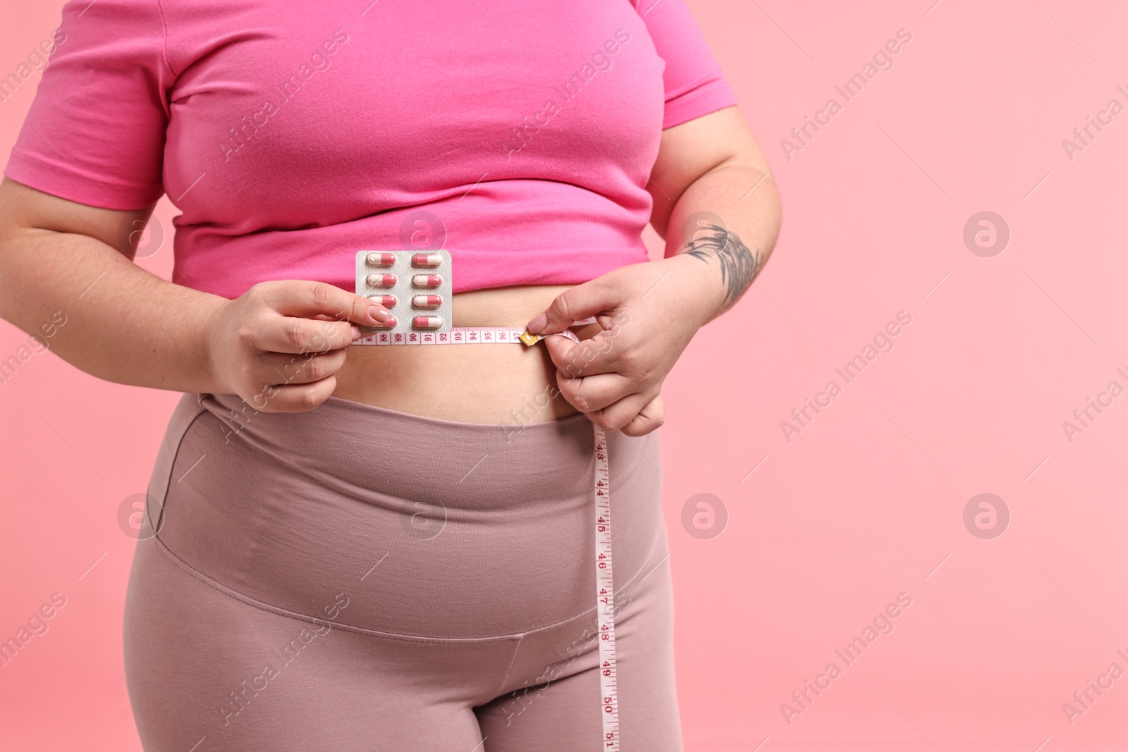 Photo of Plus size woman measuring waist with tape and holding blister of weight loss supplements on pink background, closeup. Space for text