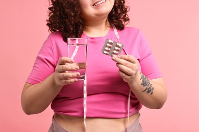 Photo of Happy plus size woman holding blister of weight loss supplements and glass of water on pink background, closeup