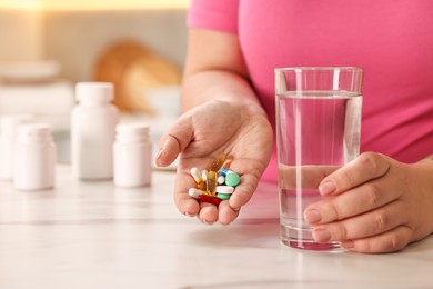 Photo of Plus size woman holding pile of weight loss supplements and glass with water at marble countertop in kitchen, closeup. Space for text