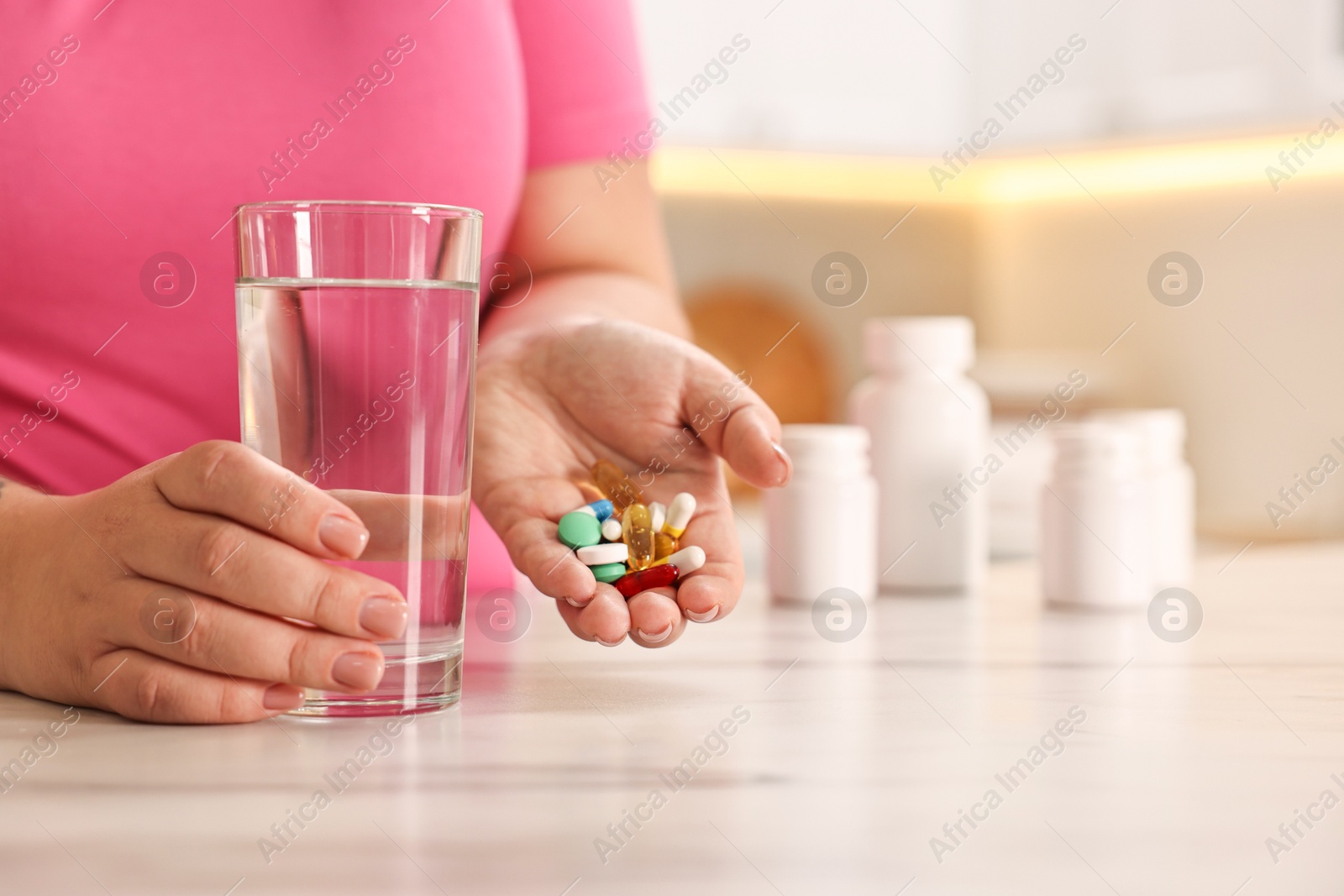 Photo of Plus size woman holding pile of weight loss supplements and glass with water at marble countertop in kitchen, closeup. Space for text