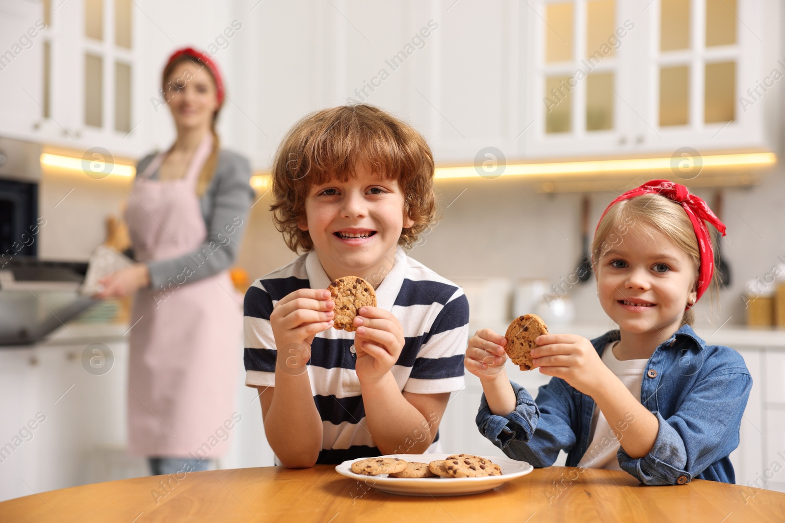 Photo of Happy kids eating tasty cookies while their mother baking in kitchen, selective focus