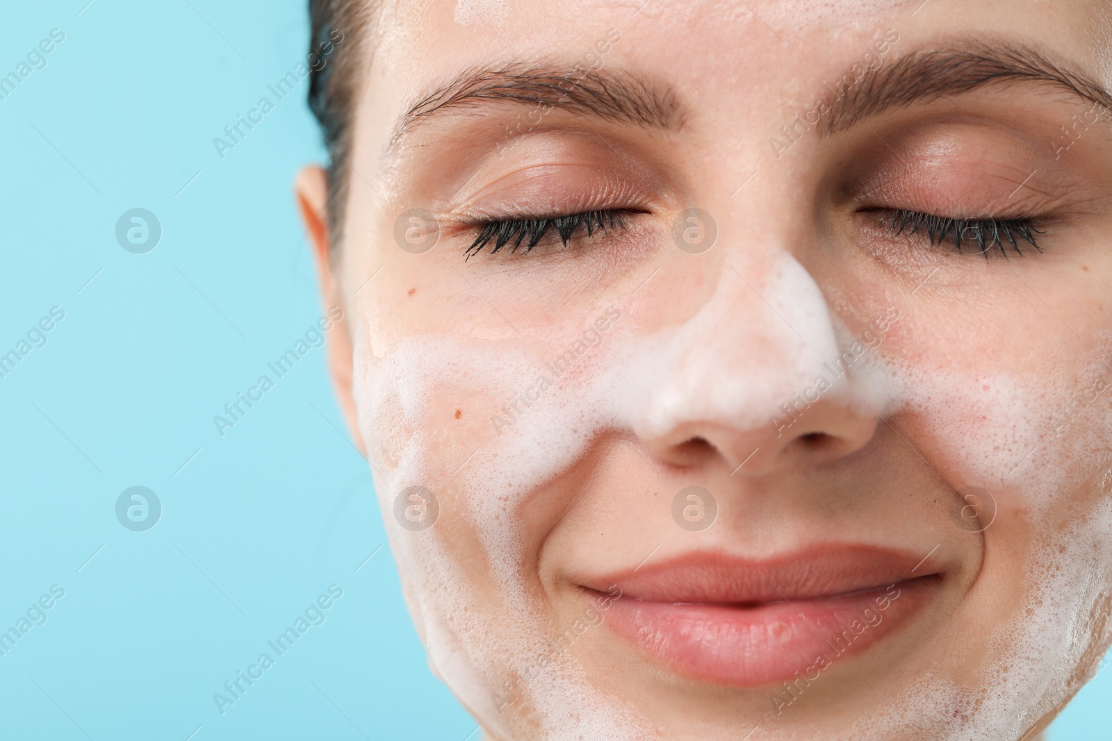 Photo of Attractive woman washing her face with cleansing foam on light blue background, closeup. Space for text